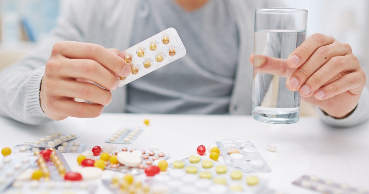 Person hält eine Blisterpackung mit Tabletten in der einen Hand und ein Glas Wasser in der anderen Hand, mit verschiedenen Medikamenten auf dem Tisch.